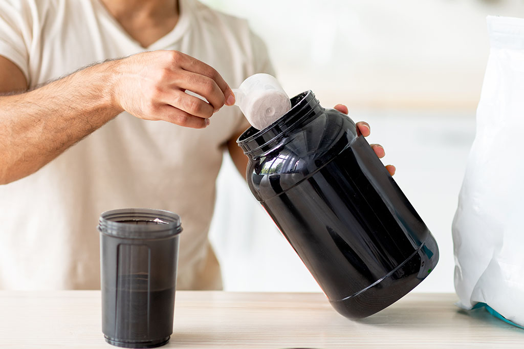 Unrecognizable young sporty guy making protein shake at table in kitchen, closeup. Cropped view of millennial bodybuilder using sports nutrition supplement, preparing cocktail indoors